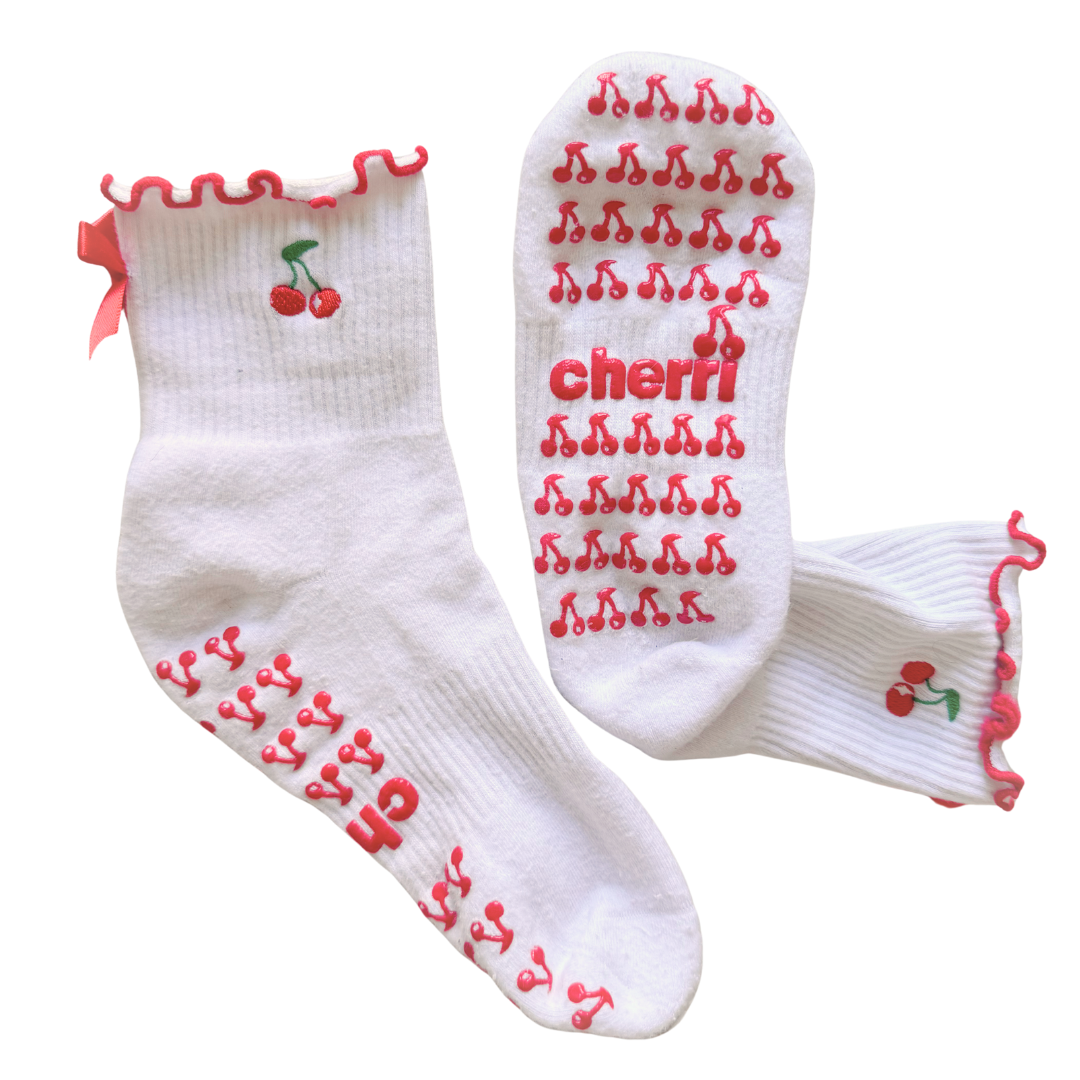 Cherri Ruffle Pilates Grip Socks - flat lay showing white crew socks with red cherry print, cherri logo, pink ruffled lettuce-trim edge, and satin bow detail