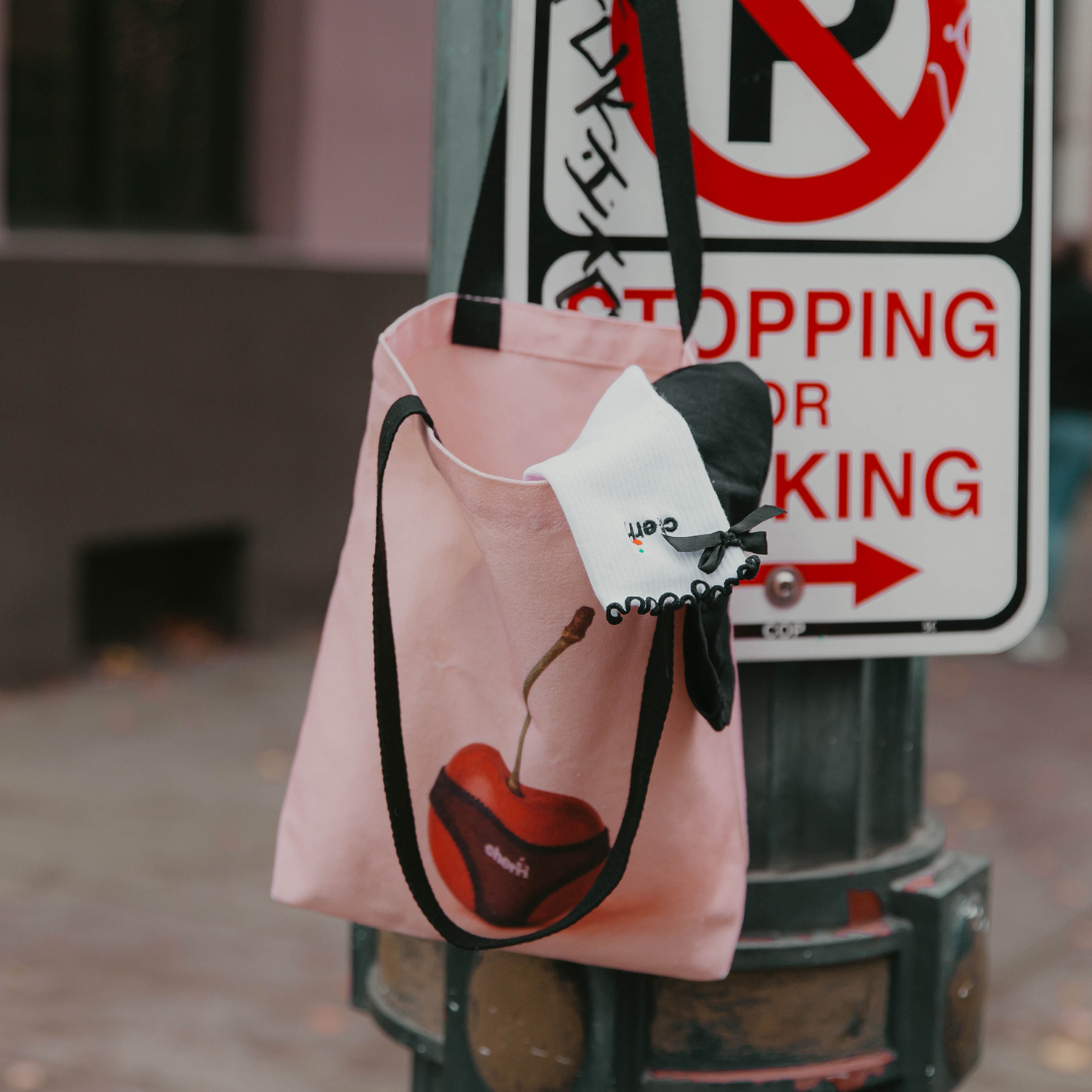 Cherri Tote Bag lifestyle shot - pink canvas tote with cherry design hanging on street sign post with Cherri socks peeking out