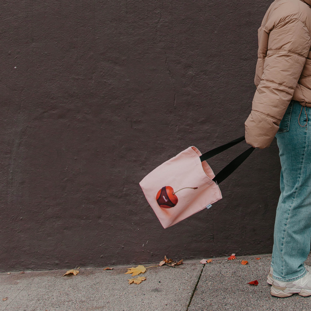 Cherri Tote Bag on model - person carrying pink canvas tote with cherry design against dark wall wearing beige puffer jacket