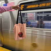 Cherri Tote Bag lifestyle shot - person holding pink canvas tote with cherry-in-underwear design on NYC subway platform with F train passing